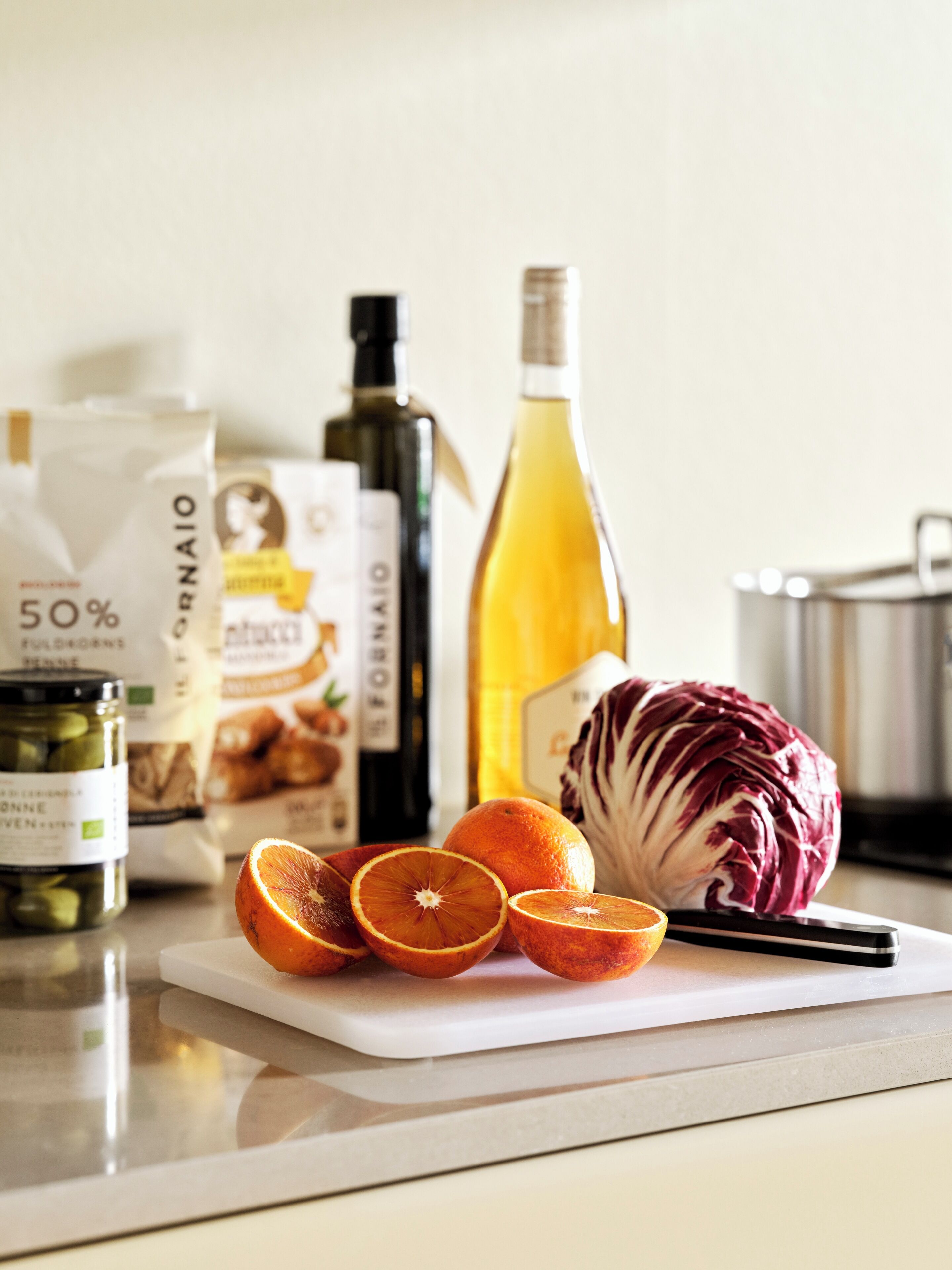Una encimera de cocina con tabla de cortar, naranjas, un repollo y botellas de aceite de oliva y vino.