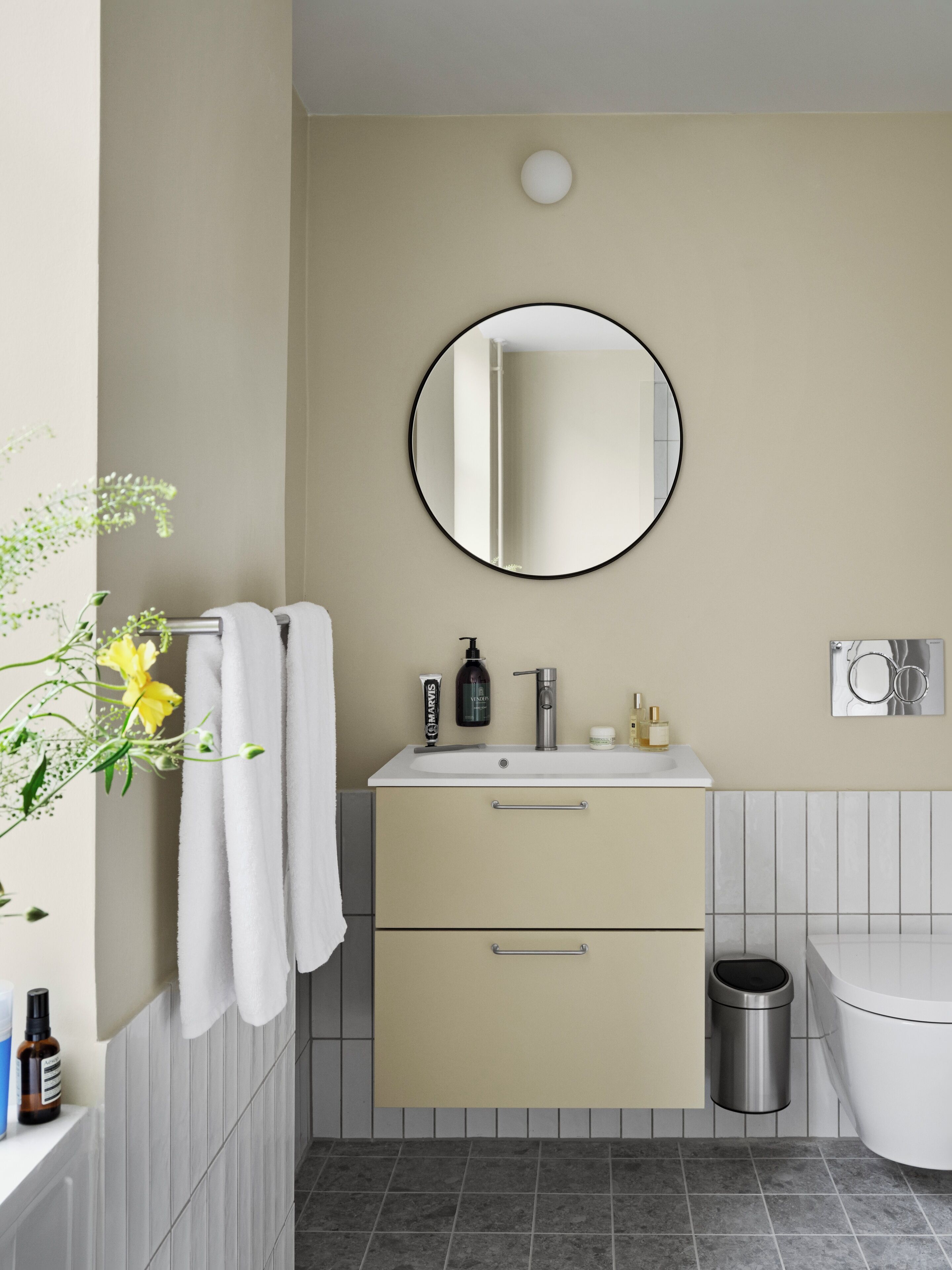 A modern bathroom with a sink, mirror, and toilet. There are towels hanging on a rack, a trash can, and toiletries on the sink.