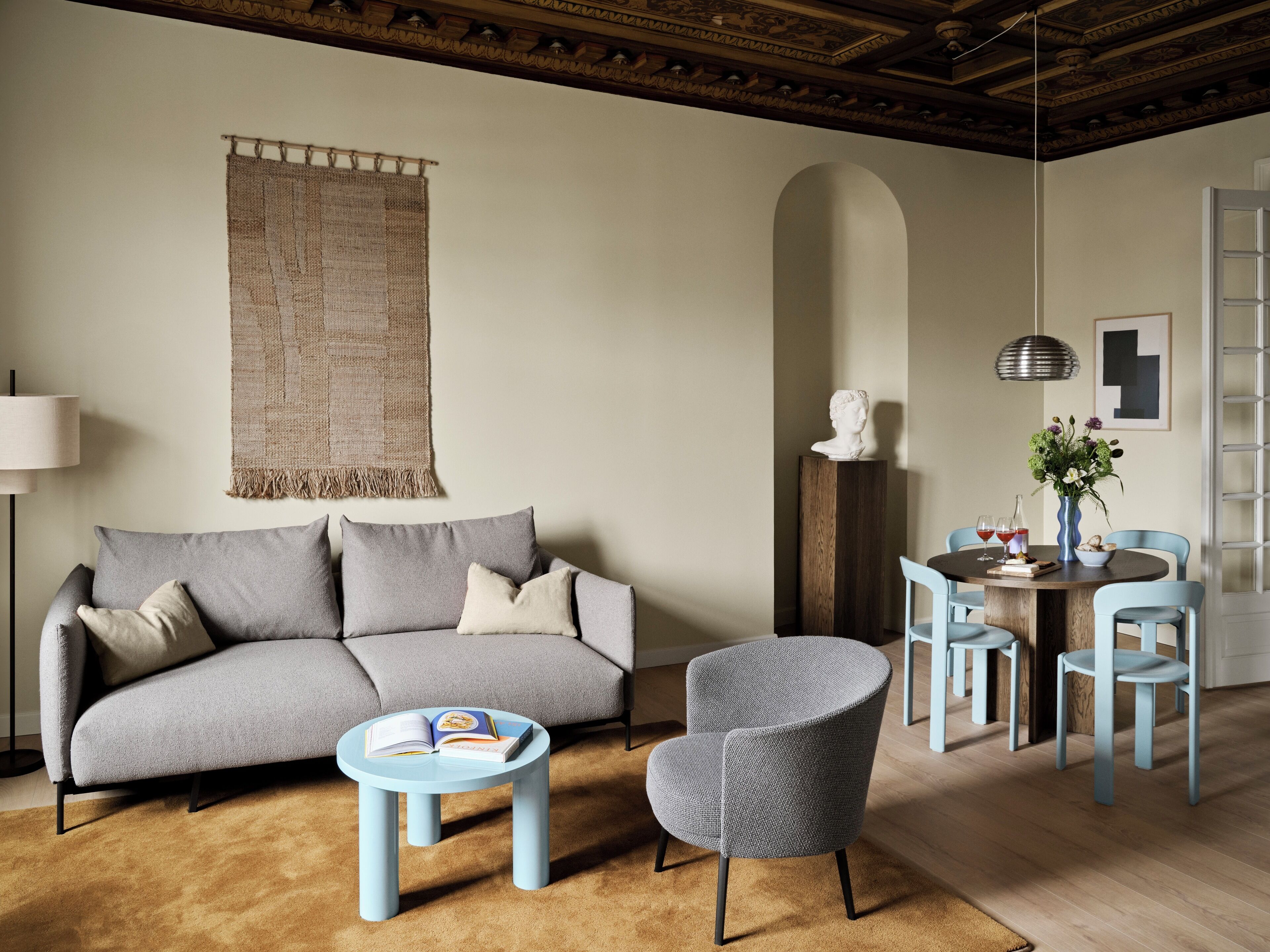 A living room with a grey sofa, a round coffee table, and a blue armchair. There is a dining area with a round table and blue chairs. A tapestry hangs on the wall, and a bust is displayed on a pedestal.