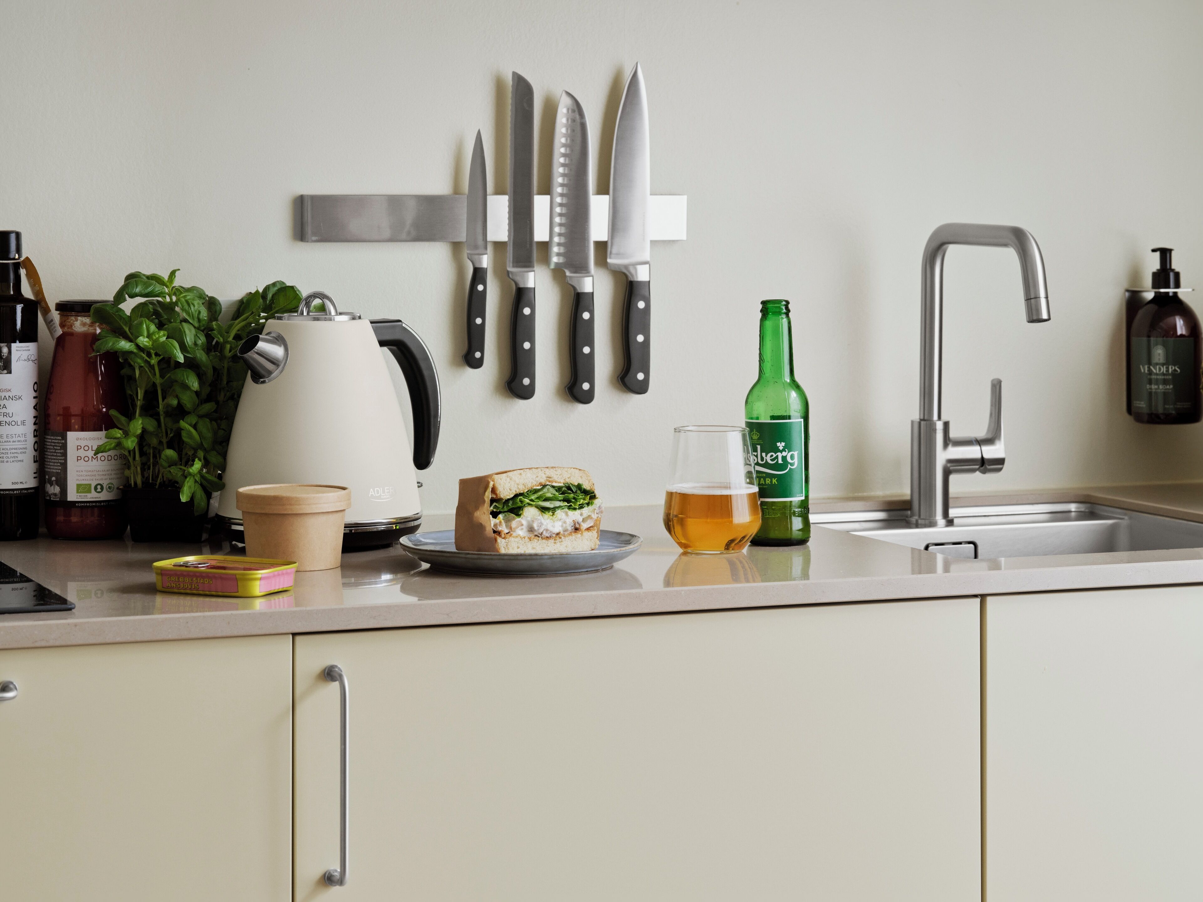 A kitchen with a sandwich on a plate, a glass of beer, a green bottle, and several knives on the wall.
