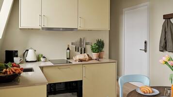 A compact kitchen with a built-in oven, a coffee maker, and a kettle. There is a dining area with a table, chairs, and a plate with a croissant.