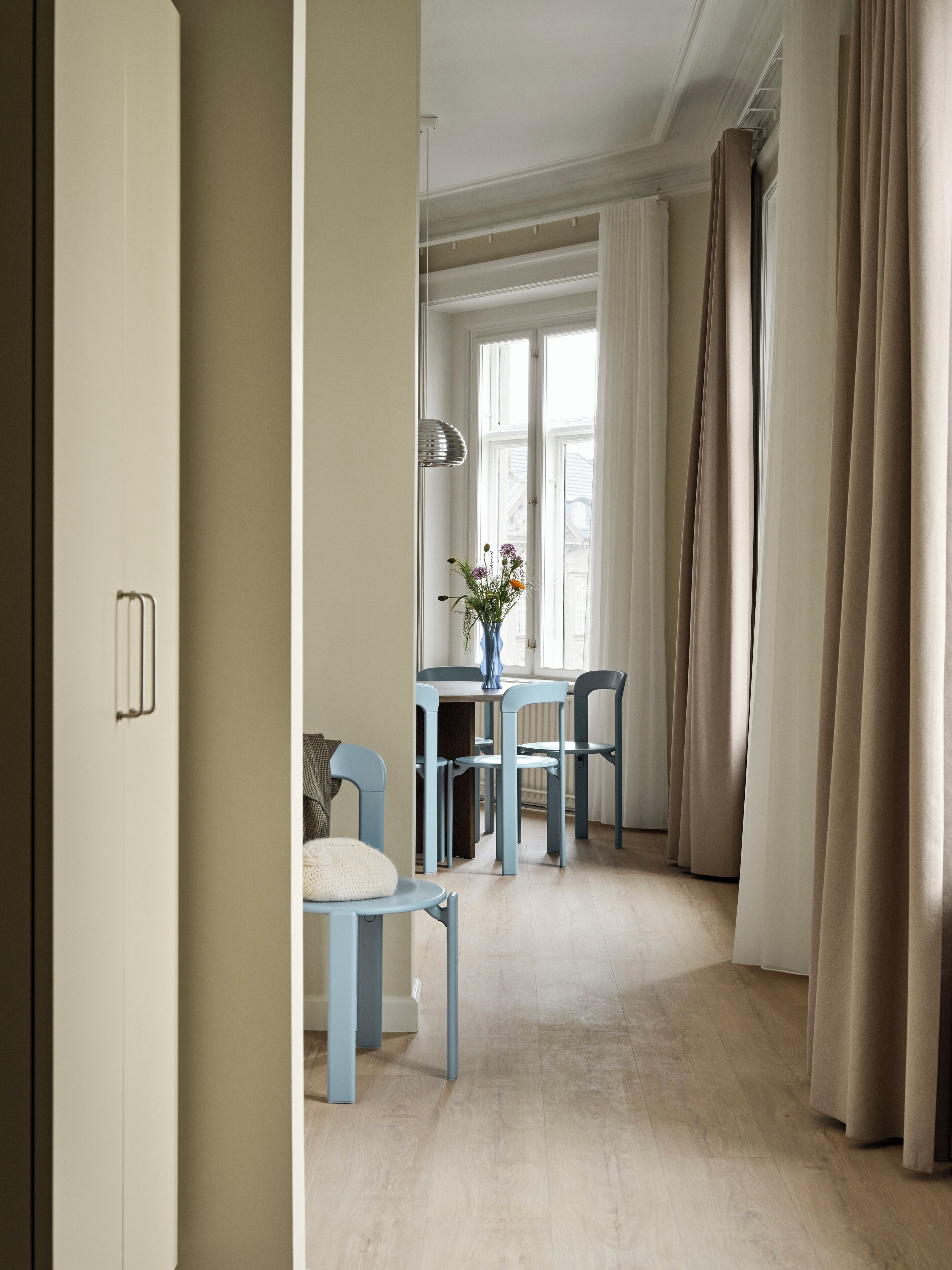 Ett rum med en matplats som inkluderar ett bord och stolar, en vas med blommor och en utsikt genom ett fönster med gardiner.