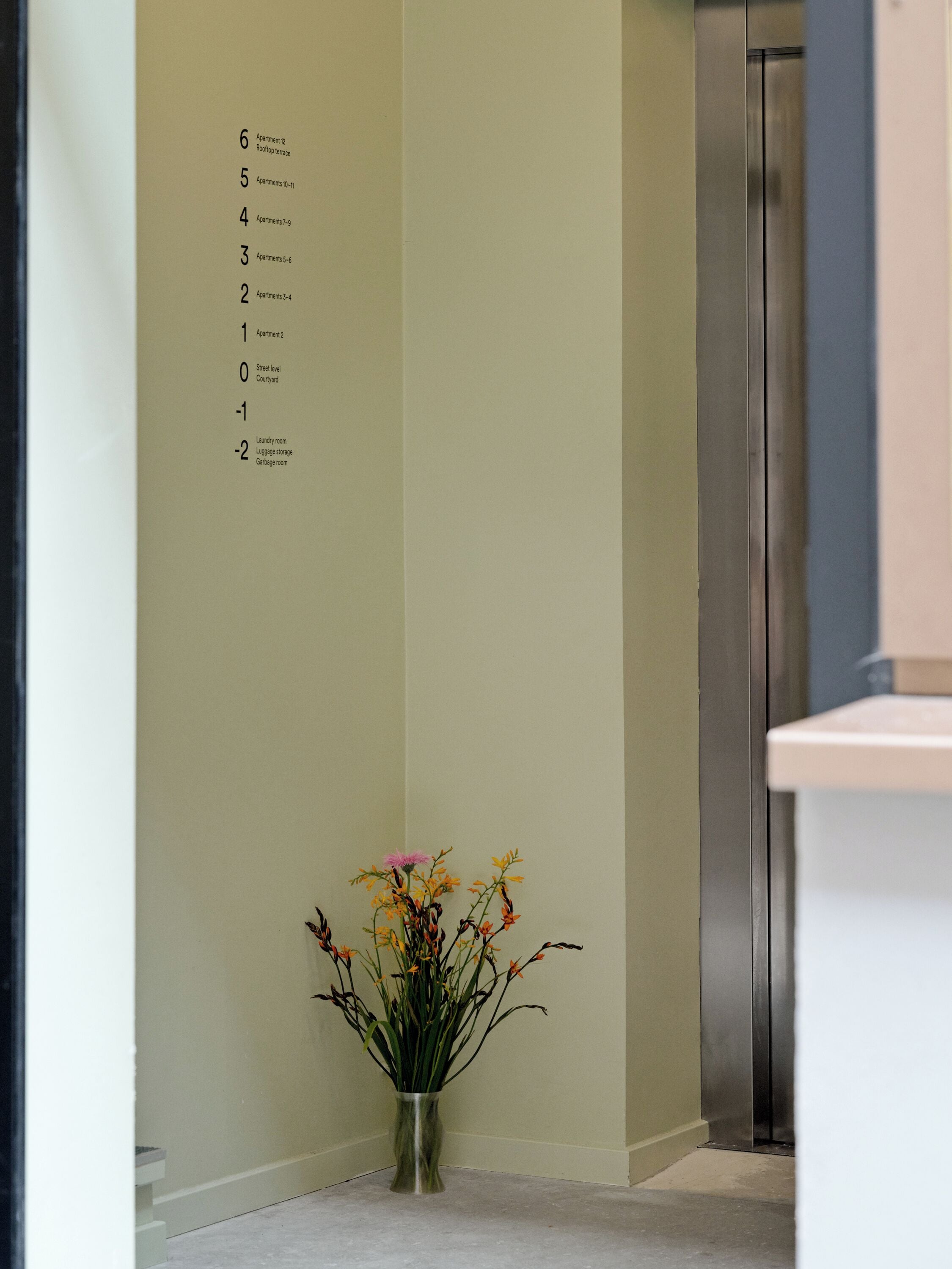 A hotel corridor with a wall-mounted directory listing rooms and facilities, a vase with flowers, and an elevator.