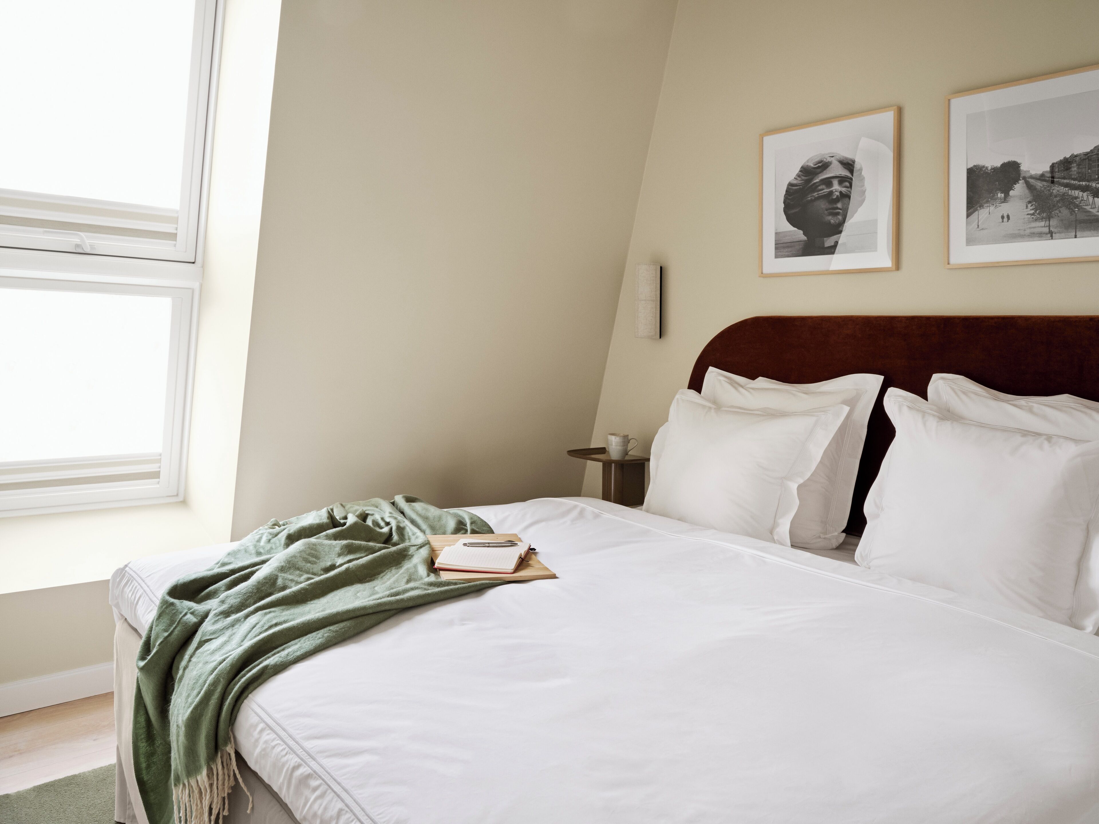 A neatly made bed with white linens, a green throw blanket, and a book on the side. There are two framed pictures on the wall above the bed.
