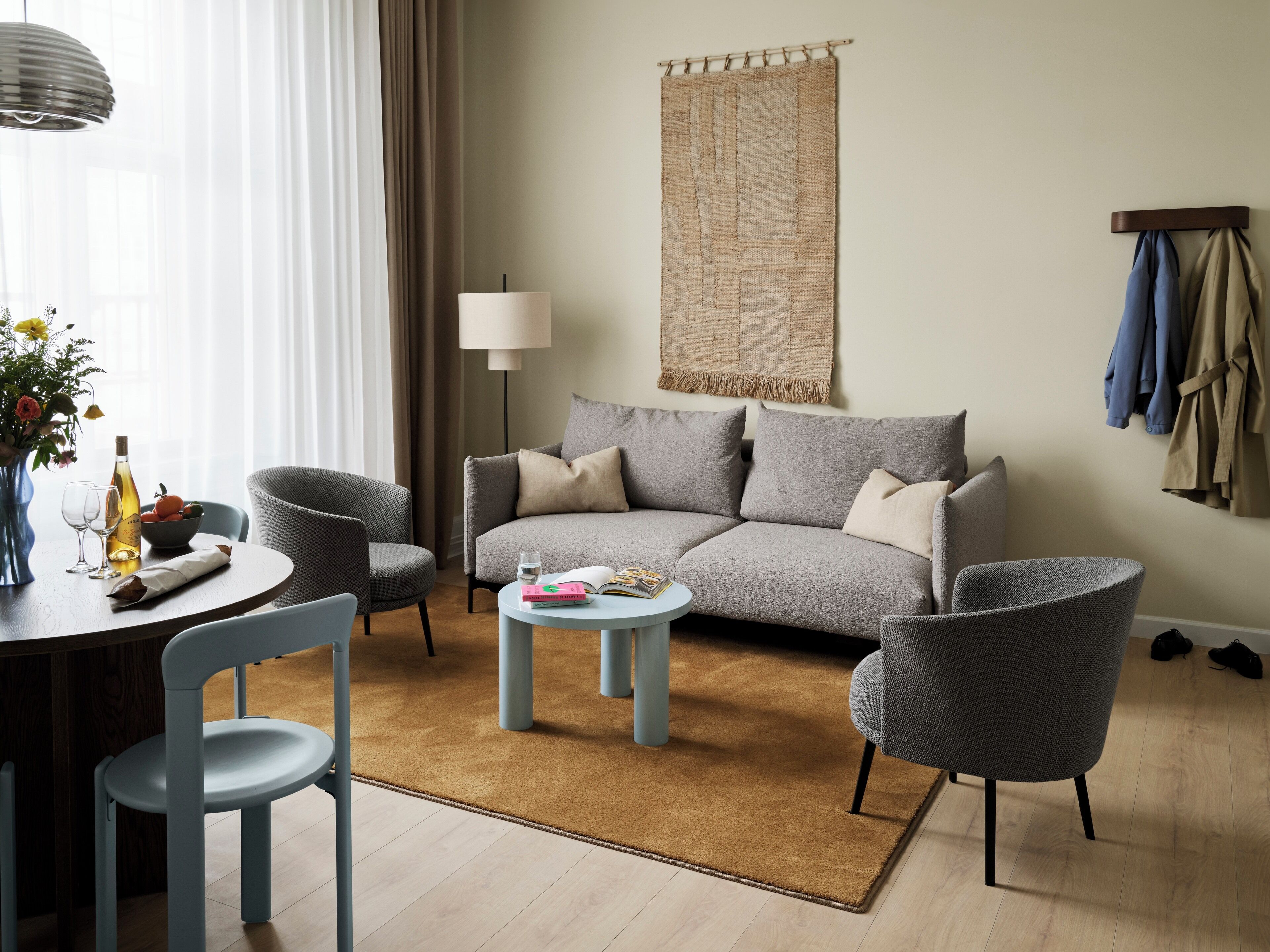 A modern living room with a sofa, two armchairs, a coffee table, and a dining table with chairs. There is a lamp, a wall tapestry, and a coat rack with clothes.