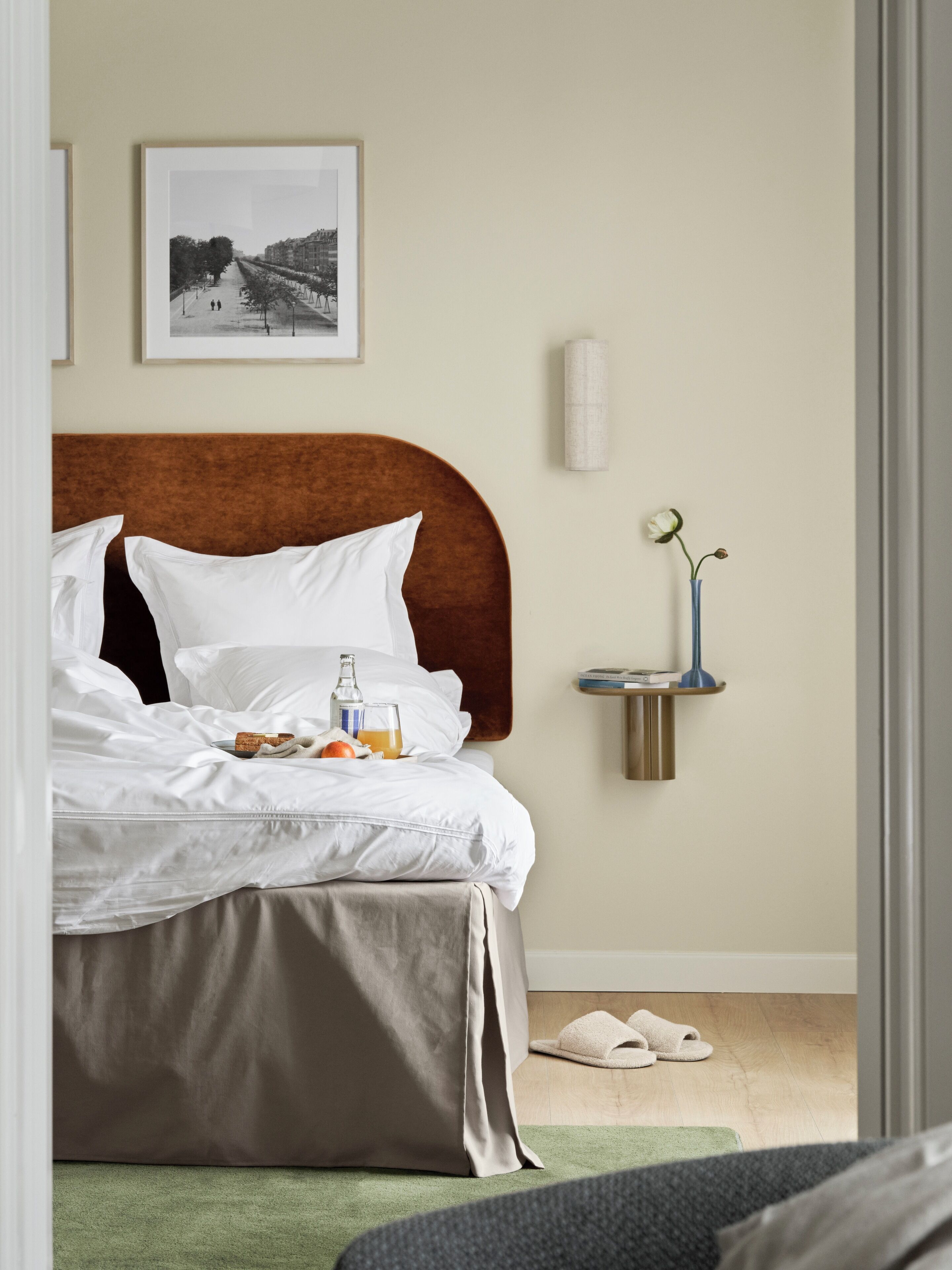 A neatly made bed with white linens, a brown headboard, and a bedside table with a vase and a lamp.