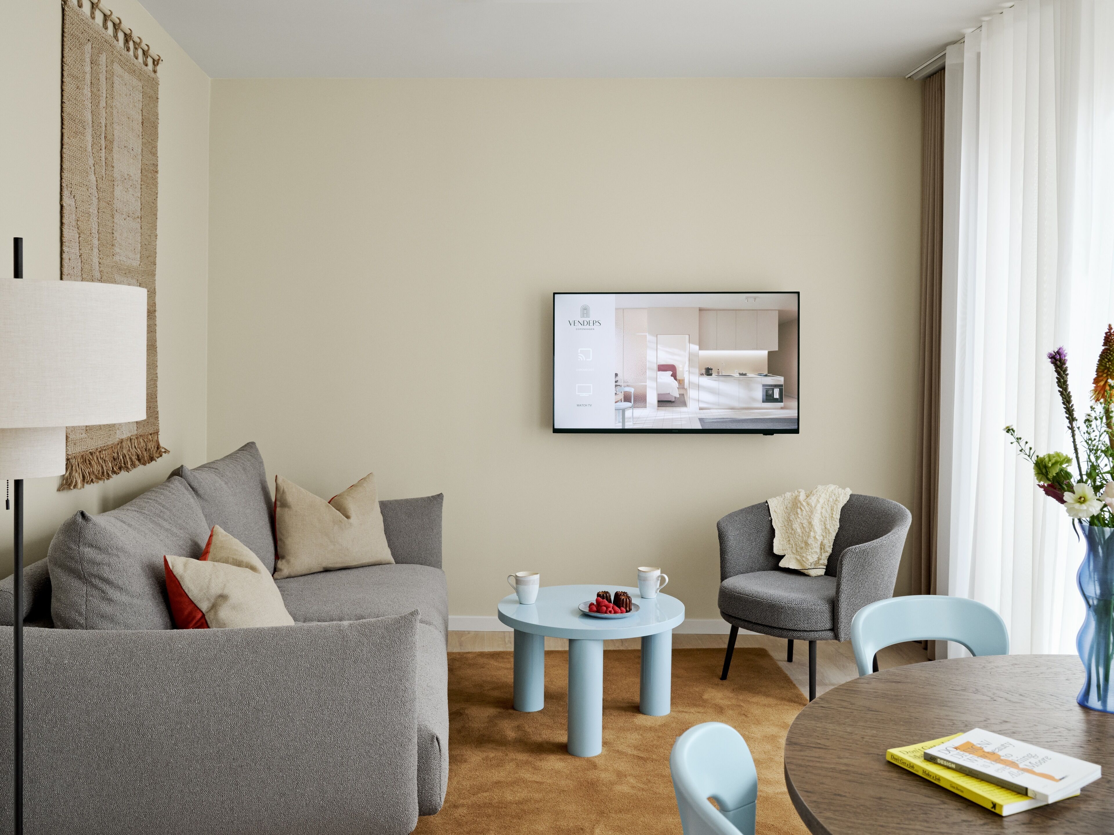 Habitació d'hotel moderna amb un sofà gris, una taula rodona petita amb una tassa i aperitius, un armari gris, una taula de fusta rodona amb llibres i una televisió que mostra un plànol de l'espai.