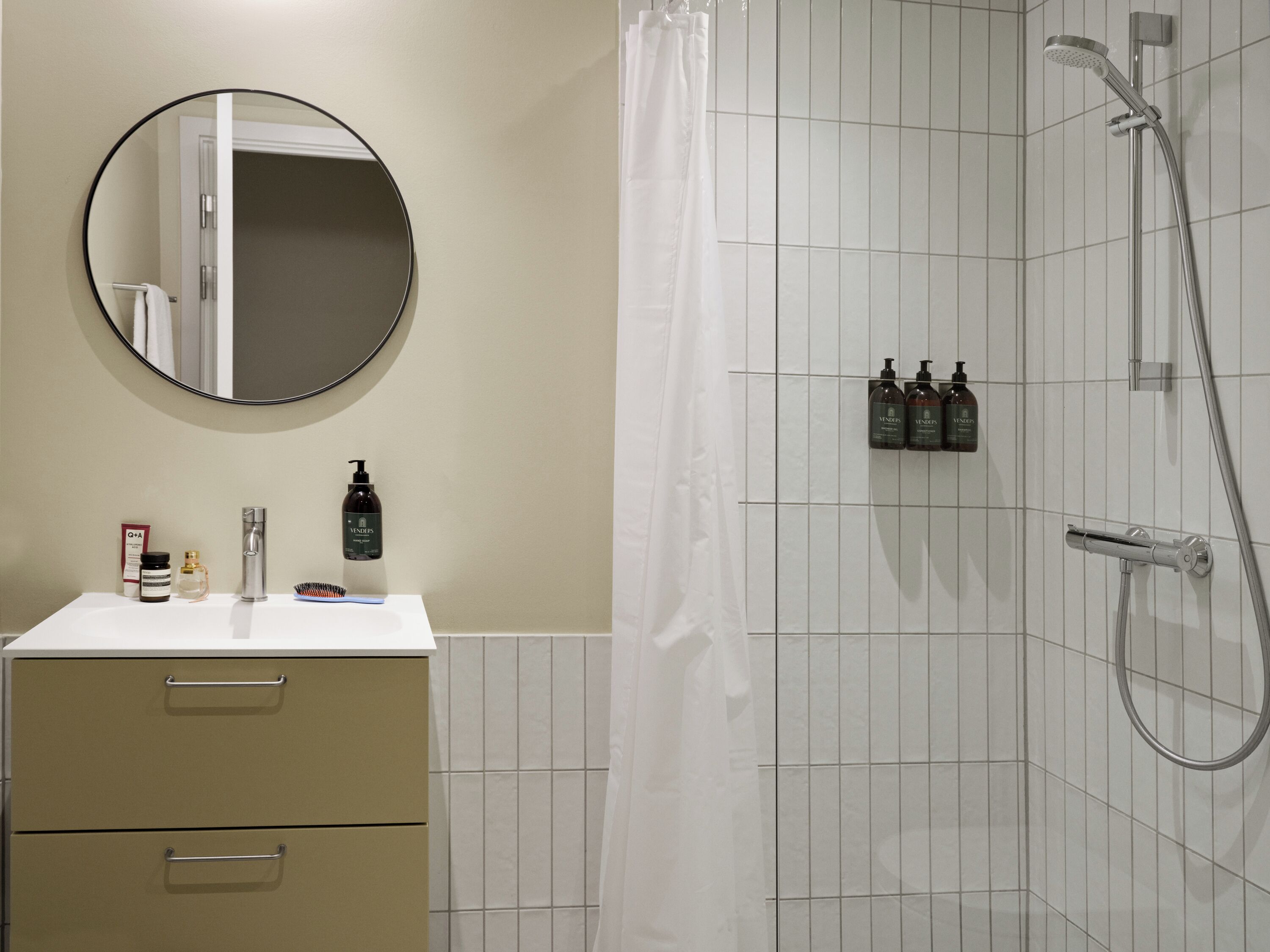 A bathroom with a white sink, mirror, and toiletries. There is a shower with a handheld showerhead and a curtain.