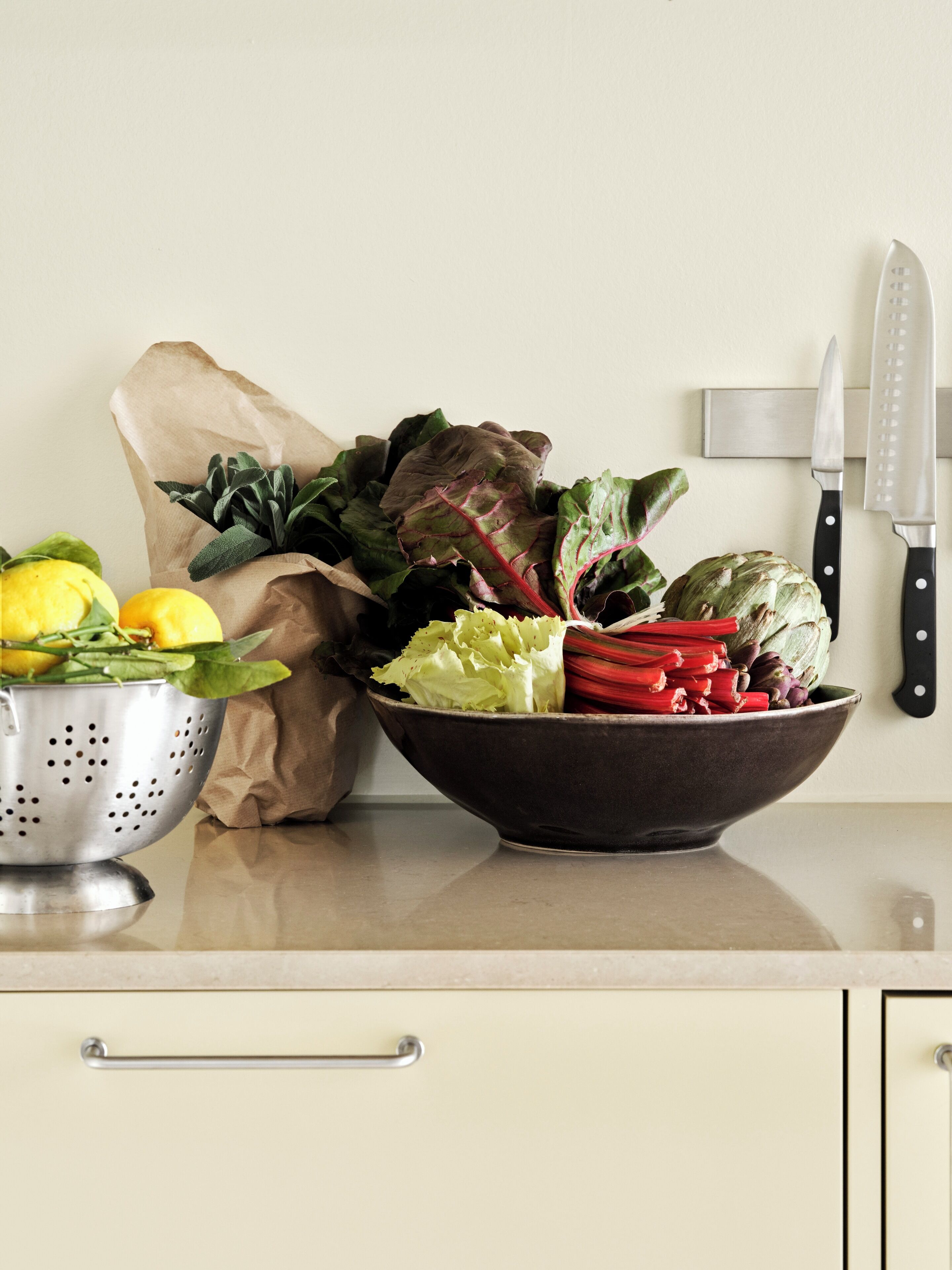 A kitchen counter with a colander, a paper bag, a bowl of mixed vegetables, and two knives.