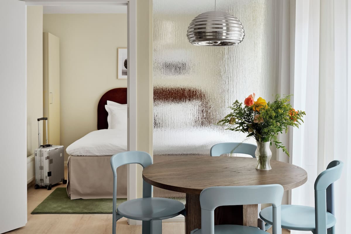 Een hotelkamer met een ronde houten tafel, vier lichtblauwe stoelen, een bed met wit beddengoed, een koffer en een vaas met bloemen.