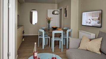 A modern living room with a sofa, a dining table with chairs, a TV mounted on the wall, and a kitchen area visible through an open door.