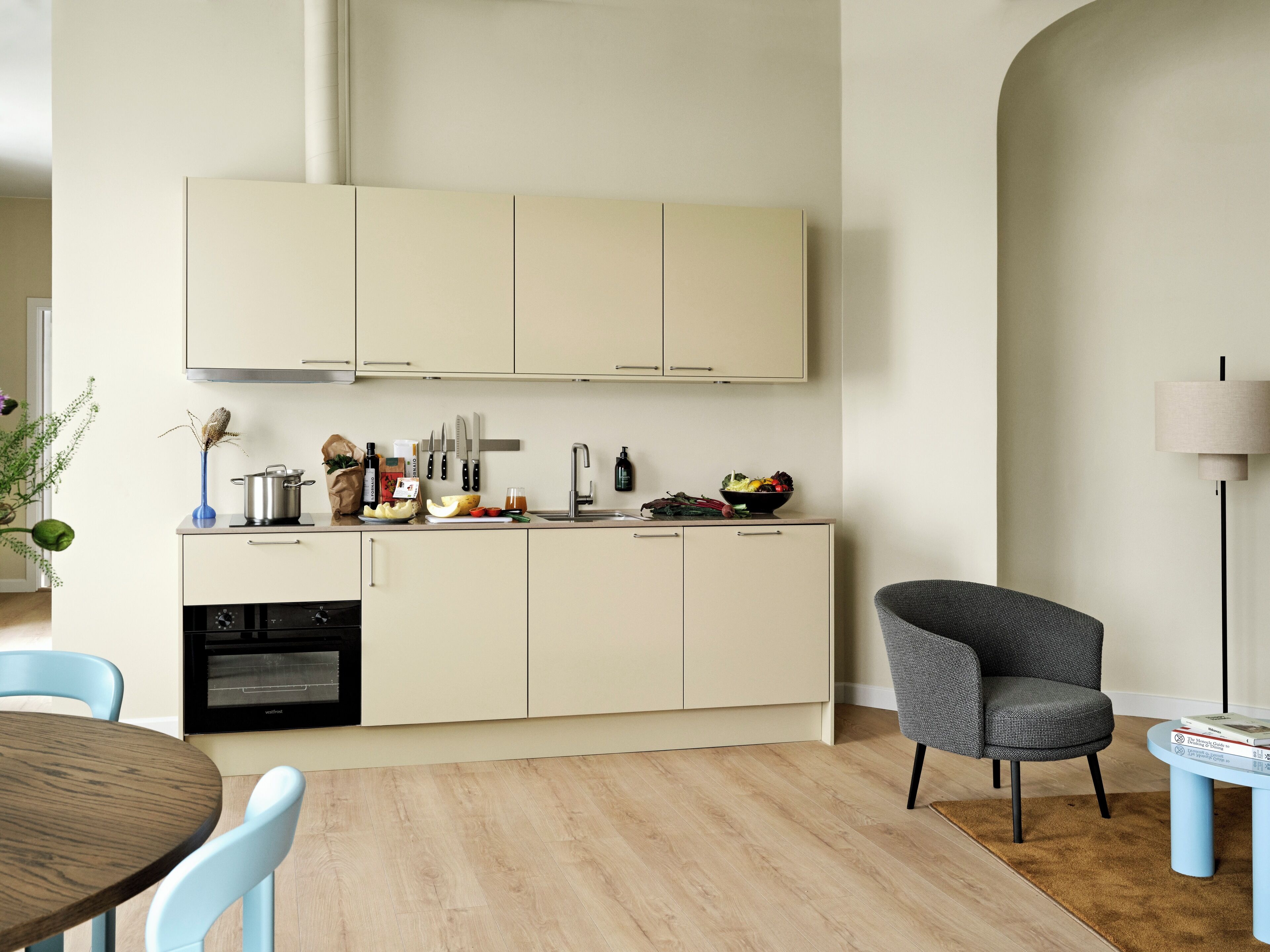 A modern kitchen with light-colored cabinets, a sink, and various kitchen items. There is a dining table with chairs and a lamp.