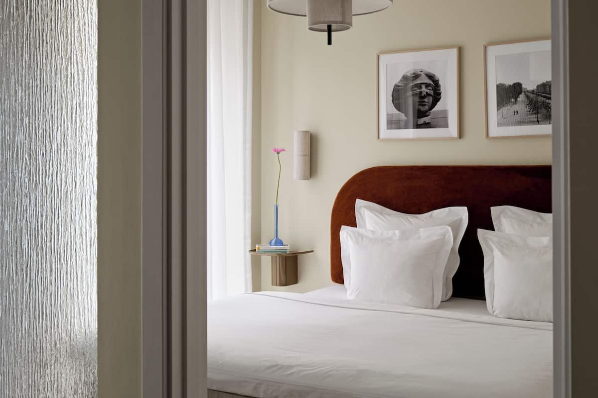 Een hotelkamer met een bed, witte kussens, een nachtkastje met een vaas, en twee foto's in lijsten aan de muur.