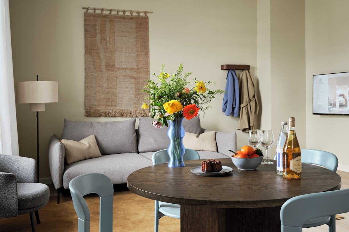 Een eethoek met een ronde tafel, stoelen, een vaas met bloemen, een schaal met fruit, wijnglazen en een fles wijn.