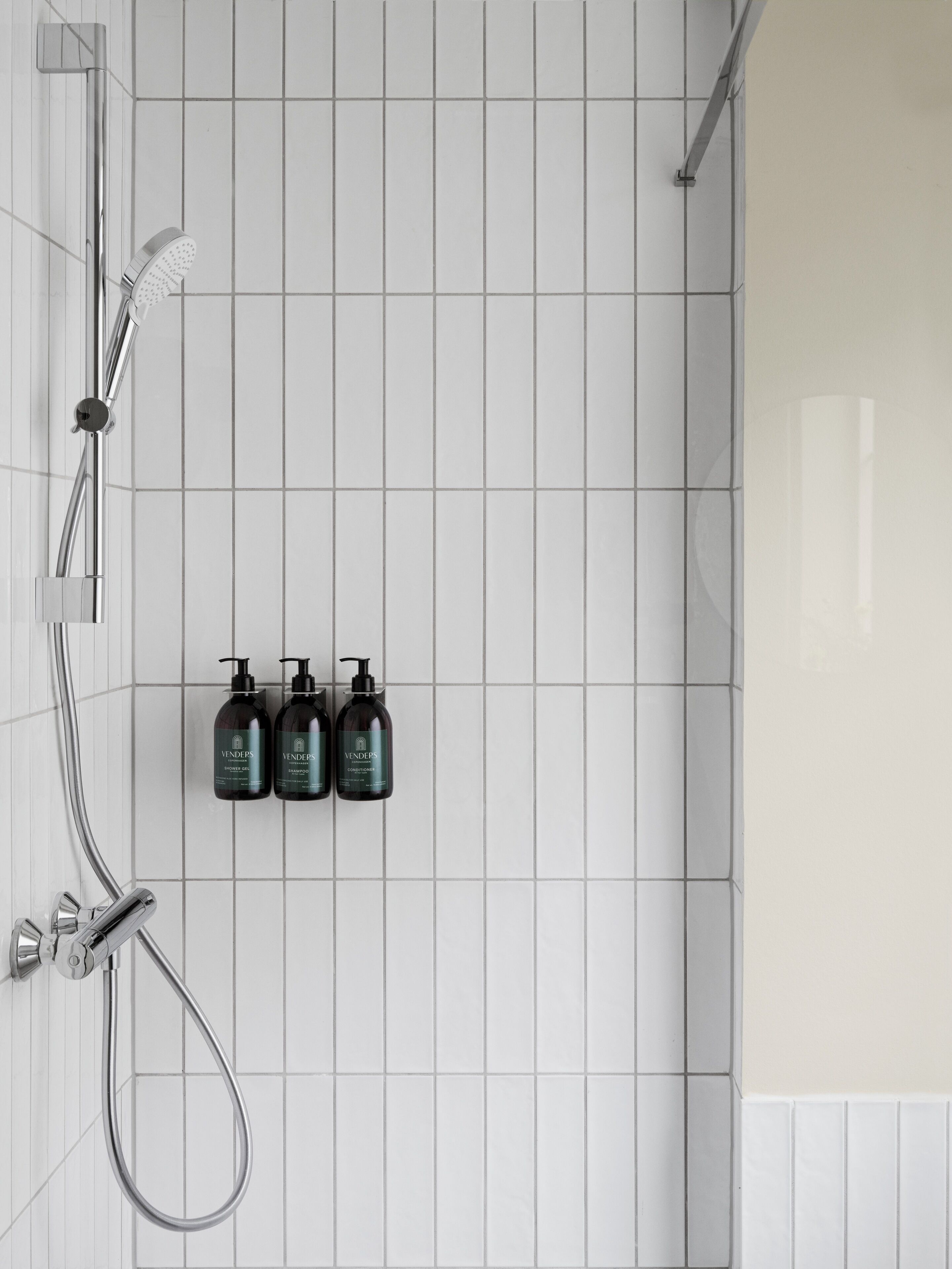 A bathroom with white tiled walls, a shower with a handheld showerhead, and three bottles of Vendeps products.