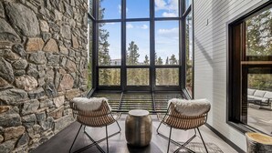 Habitación con paredes de piedra y un amplio ventanal con vistas a un bosque. Dos sillones de mimbre y una mesa de centro con textura están ubicados frente al ventanal.
