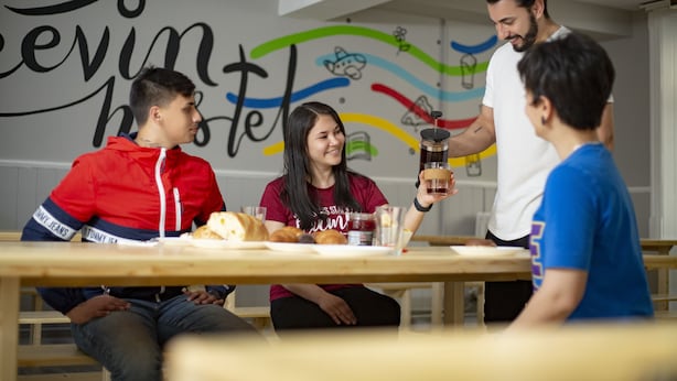 A group of people sitting at a wooden table with food and drinks, in a room with a colorful wall mural.