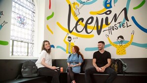 Three people sitting on a bench in a hostel with a colorful wall mural and a window with bars.