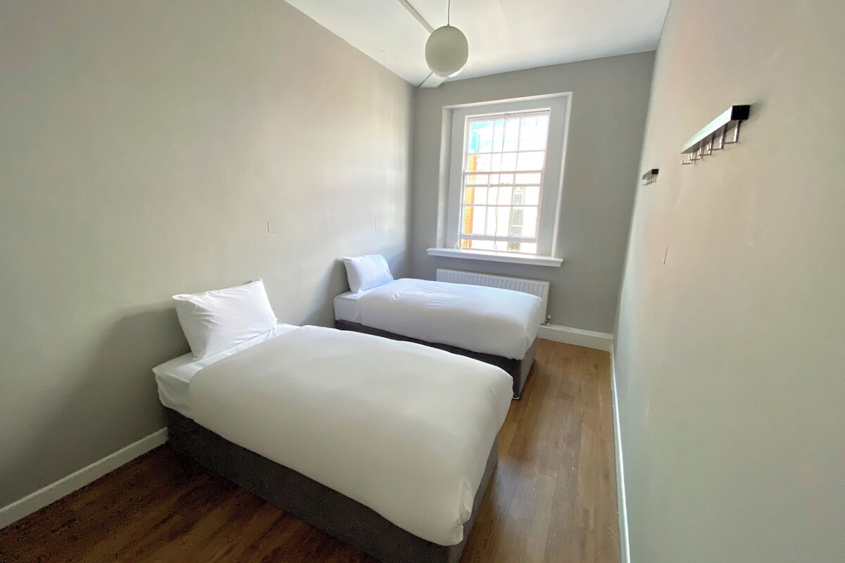 A small, bright room with two beds, a window, and a radiator.