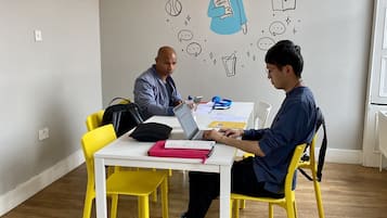 Two people seated at a white table with yellow chairs in a room with wooden flooring and a light-colored wall.
