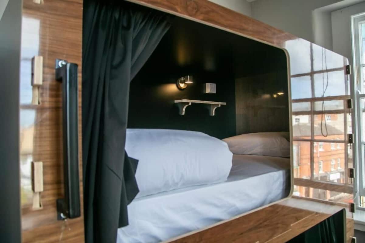 Un lit superposé avec une structure en bois, un rideau noir, une petite étagère avec une lampe et une fenêtre donnant sur des immeubles.