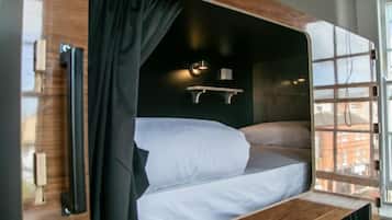 Una cama litera con estructura de madera, cortina negra, una pequeña estantería con luz y una ventana con vistas a edificios.