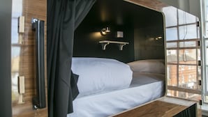 A bunk bed with a wooden frame, a bedside table, a wall-mounted light, and a window with a view of buildings. - Leevin Hostel Mountjoy (Dublin)