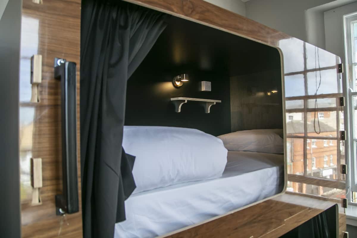 Un lit superposé avec une structure en bois, une table de chevet, une lampe fixée au mur et une fenêtre donnant sur des immeubles.