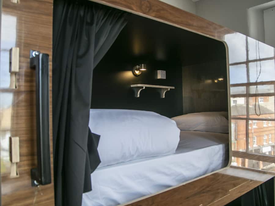Una cama litera con estructura de madera, una mesita de noche, una lámpara fijada a la pared y una ventana con vistas a edificios.