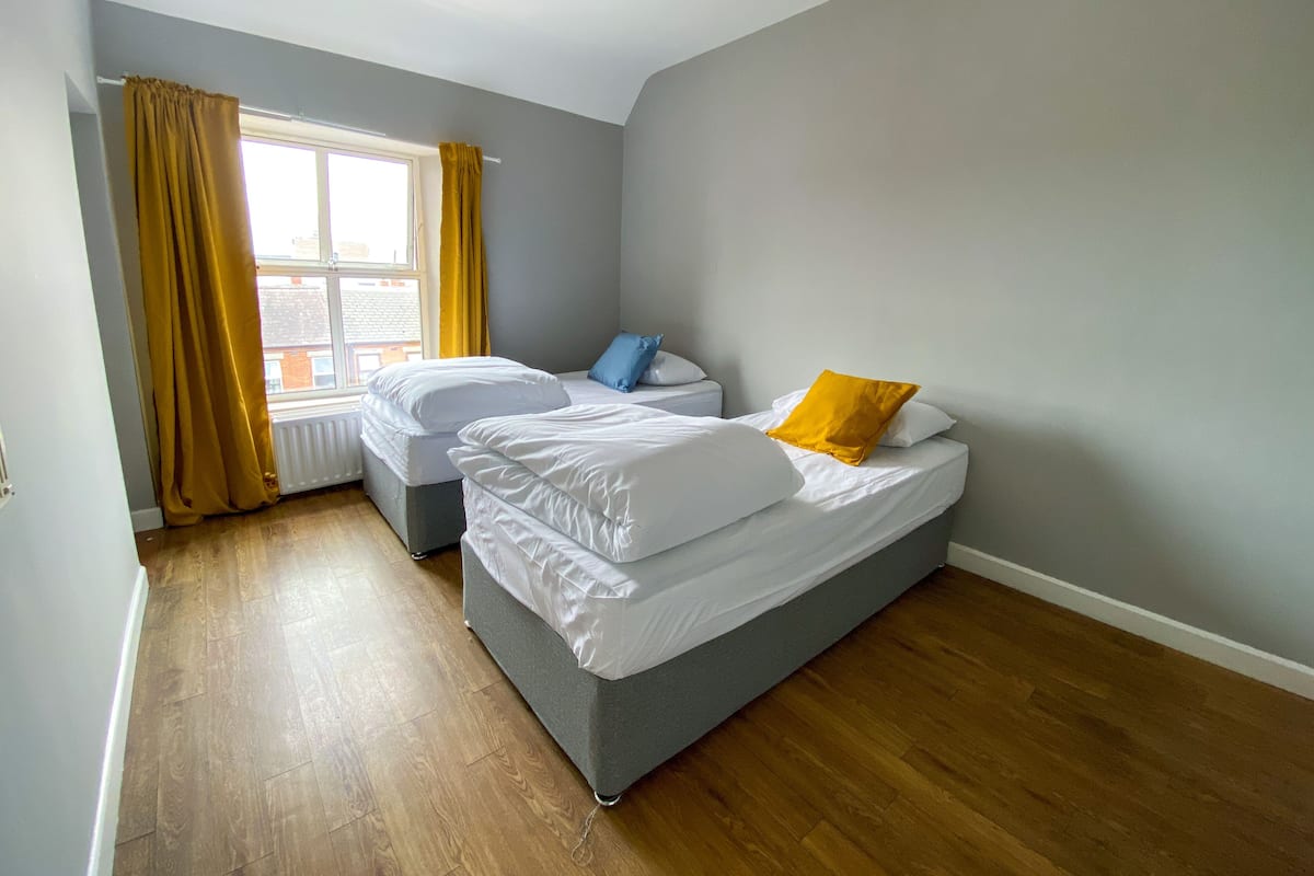 Une petite chambre moderne avec deux lits simples, une fenêtre avec des rideaux jaunes et une vue sur l’extérieur.