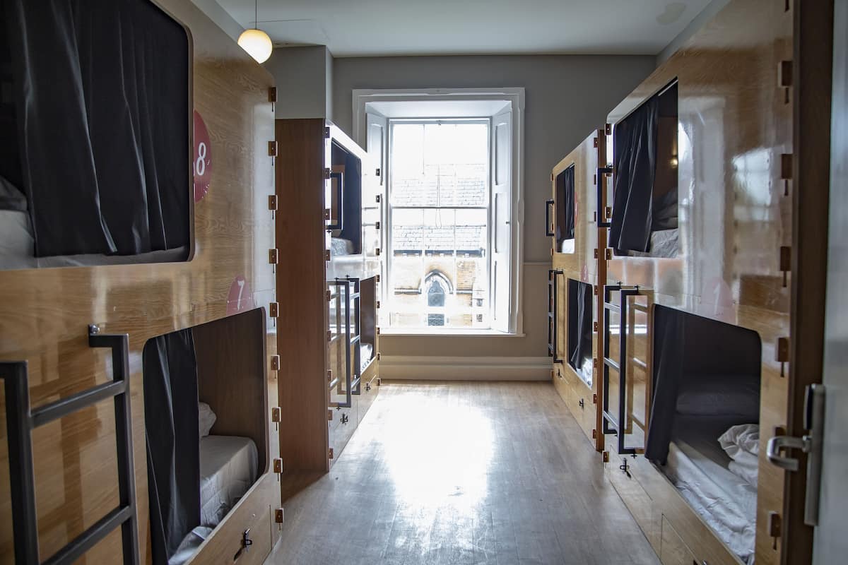 Un couloir avec des lits superposés, une fenêtre donnant sur des immeubles et une porte avec un numéro.