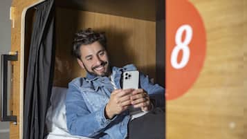 A person sitting in a wooden bunk bed room with a curtain, holding a smartphone.