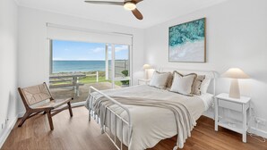 Une chambre à coucher comprenant un lit, des tables de chevet, une chaise, un ventilateur de plafond et une grande fenêtre donnant sur l’océan.