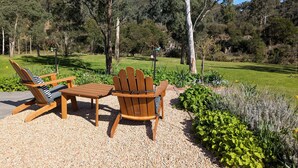 Un espacio de estar al aire libre con madera, con dos sillas tipo Adirondack y una mesa, rodeado de grava y follaje.