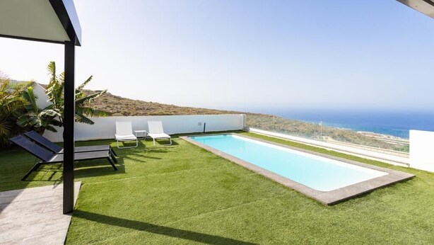 A rooftop pool with two lounge chairs and a small table, surrounded by artificial grass, overlooking a coastal landscape.