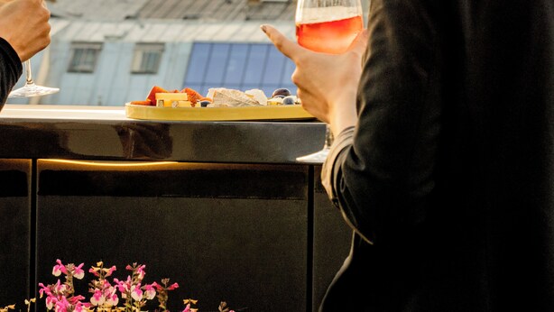 A person holding a glass of wine, another glass with a different beverage, and a plate with food items on a balcony overlooking a cityscape with a tower. - Hôtel Pilgrim (Paris)