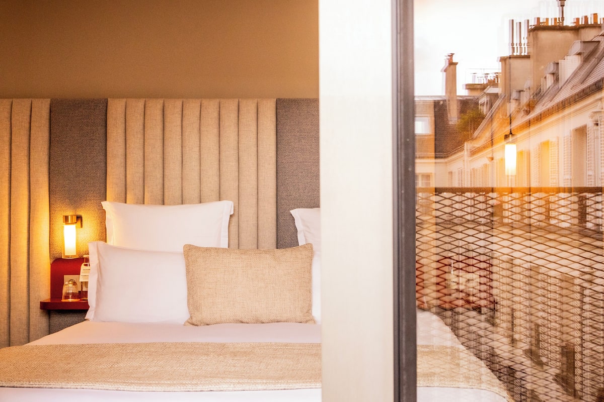 Une chambre d’hôtel avec un lit, des oreillers, une table de chevet avec une lampe et une vue sur le paysage urbain par la fenêtre.