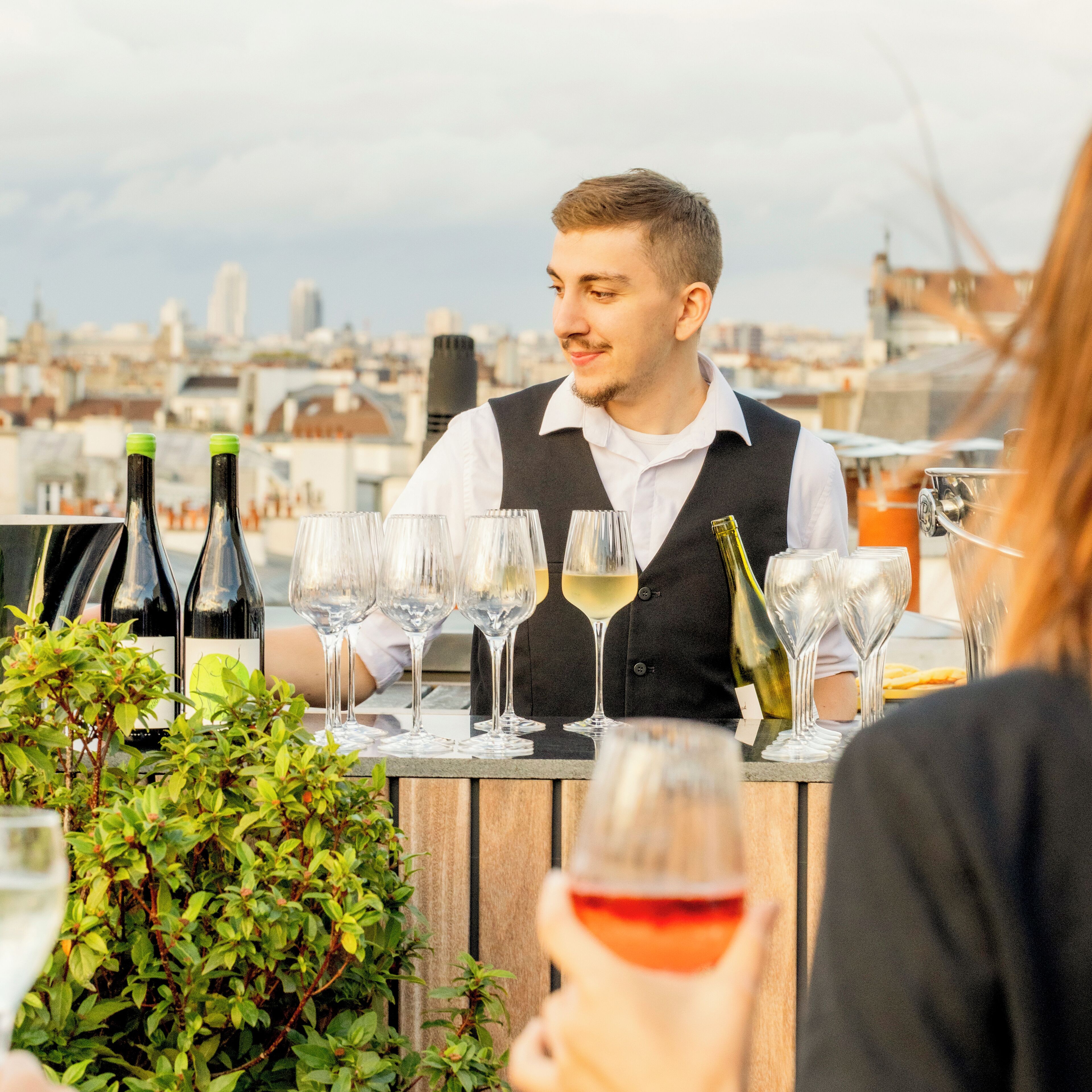 Una persona sentada a una mesa con botellas y copas de vino, con vistas a un paisaje urbano.