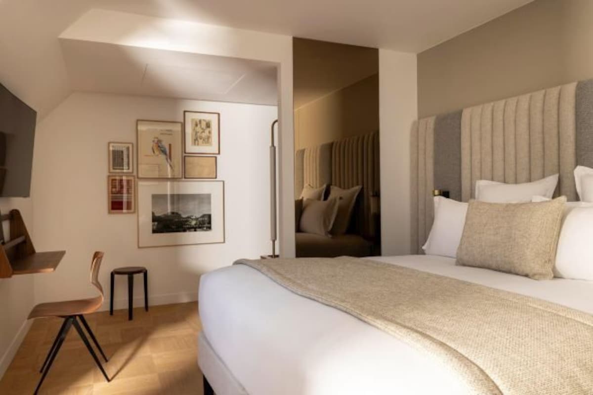 A modern hotel room with a large bed, a desk, and a chair. There are framed pictures on the wall and a lamp beside the bed.
