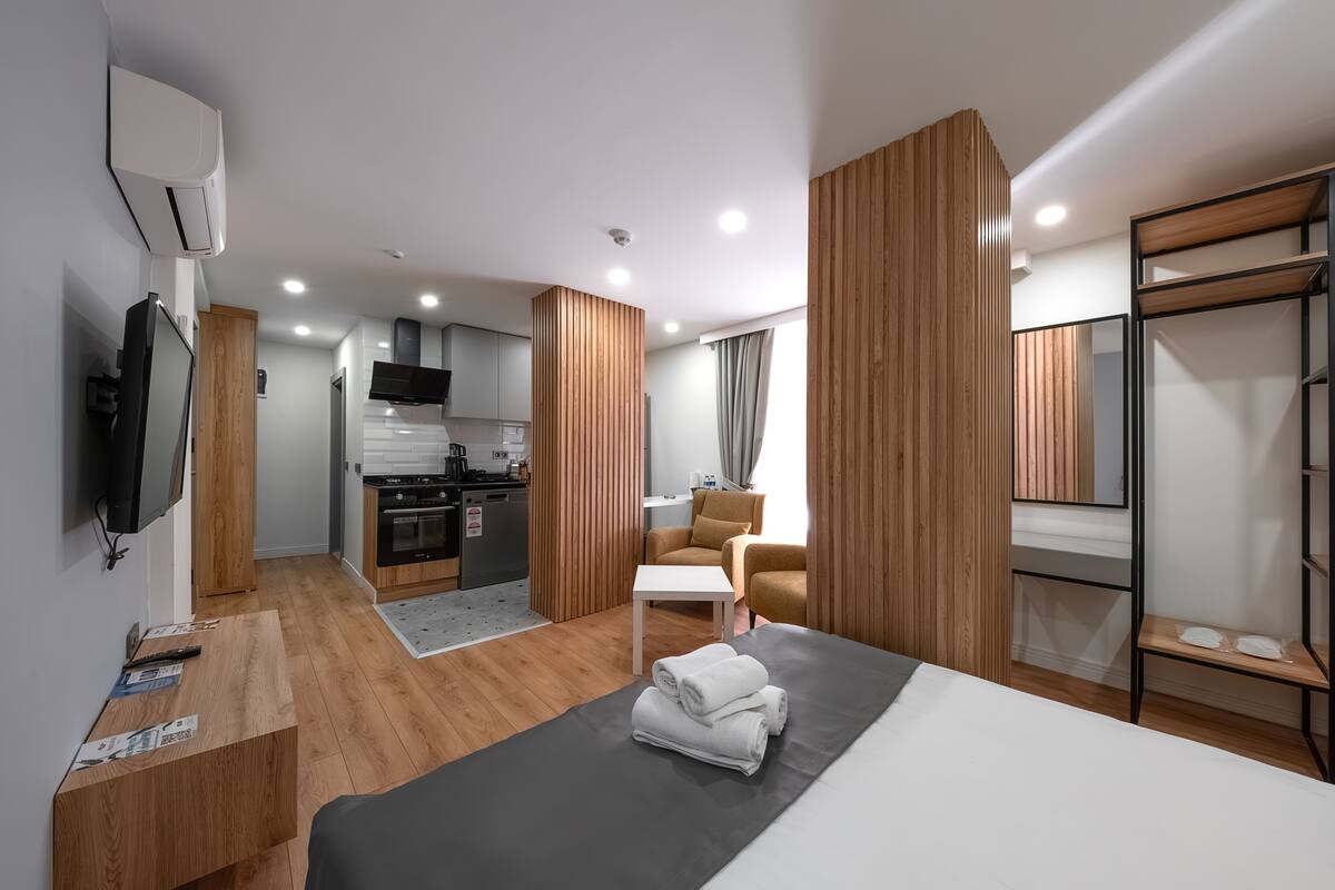 A modern hotel room with a bed, a small desk, a TV, a kitchenette, and a seating area.