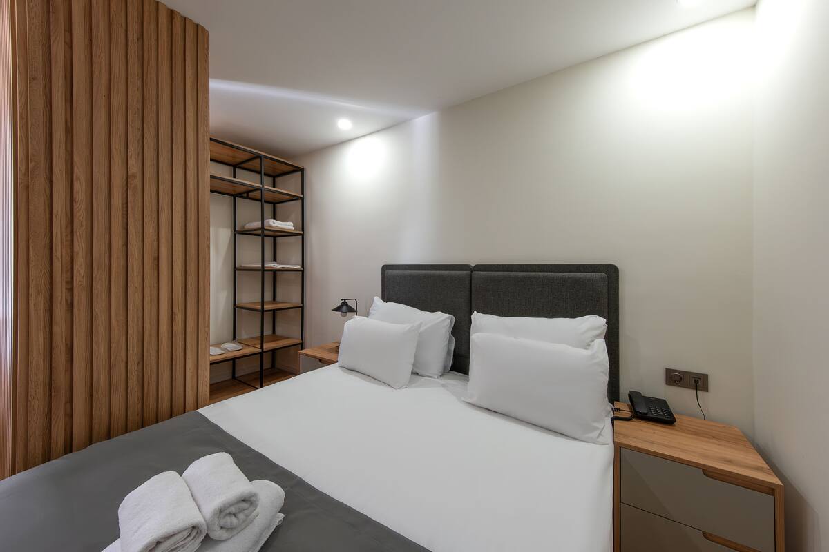 A neatly made bed with white linens, a bedside table with a phone, and a wooden wardrobe.