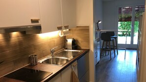 A modern kitchen with a stainless steel sink, a built-in stove, and a wooden backsplash. There is a small island with a stool and a bar area visible through a glass door. - Large Studio Near Epfl And Lausanne City Center (Ecublens)