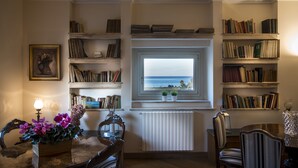 A cozy room with a view of the sea through a window, a bookshelf filled with books, a dining table with chairs, and a decorative vase with flowers. - Residence Torremuzza - Charming House In The Heart Of Palermo with lovely view (Palermo)