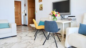 Ein modernes Wohnzimmer mit einem weißen Sofa, einem gelben Sessel und einem blauen Sessel. Ein Flachbildfernseher steht auf einem weißen Tisch, eine Vase mit Blumen und ein kleiner Tisch mit elektronischen Geräten sind ebenfalls vorhanden.