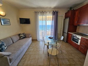 A compact living space with a sofa, a glass dining table, a kitchen with a refrigerator and stove, and a TV mounted on the wall.