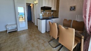 Una cocina compacta con armarios blancos, una cafetera y un lavamanos. Hay dos sillas y una mesita. Una puerta da a un balcón con vistas al mar.