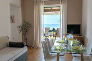 A living room with a sofa, a dining table set with glassware, a television, and a view of the sea.
