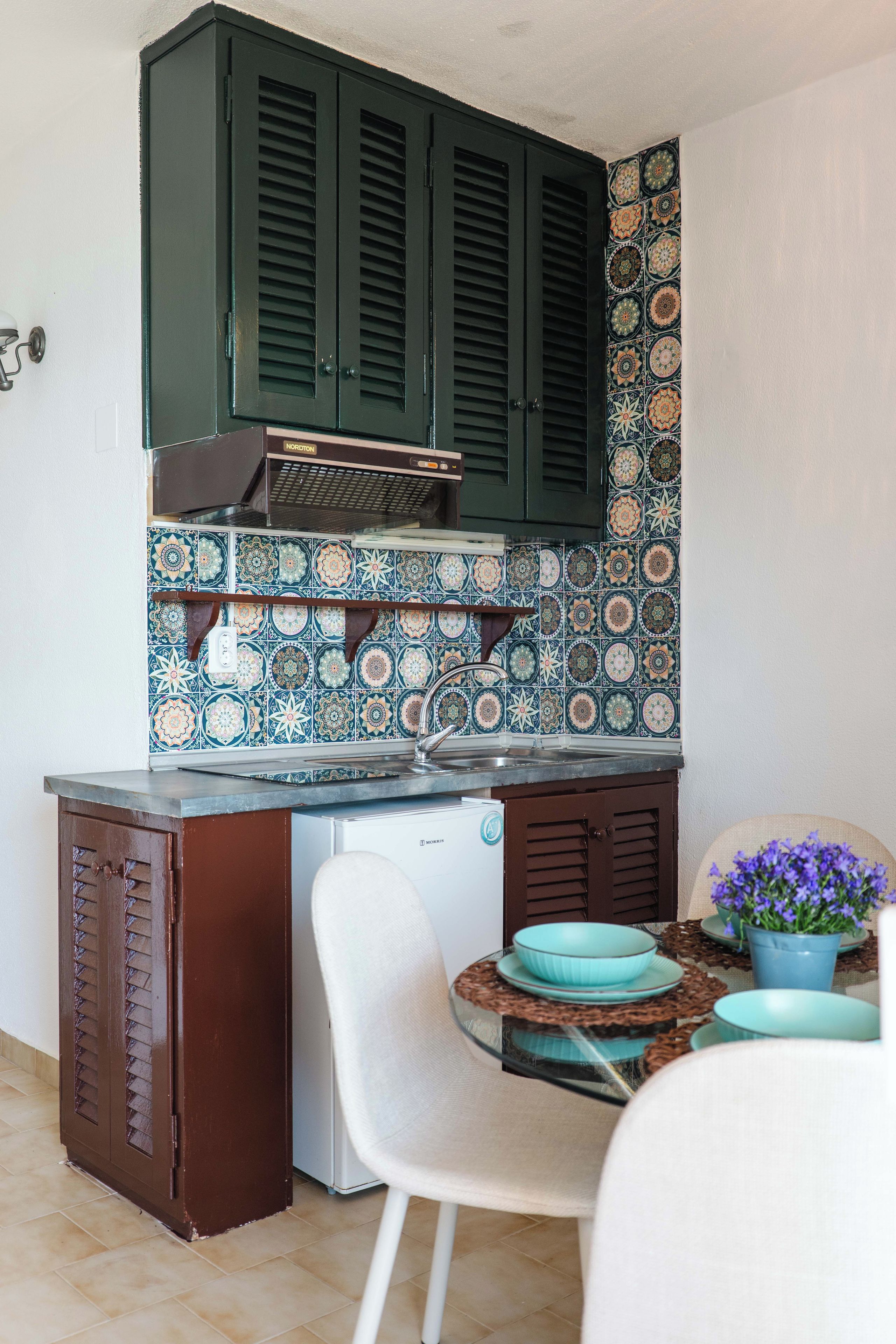 Una cucina compatta con mobili in legno scuro, un pannello decorativo dietro il piano cottura e un piccolo spazio pranzo con una sedia bianca e un tavolo.