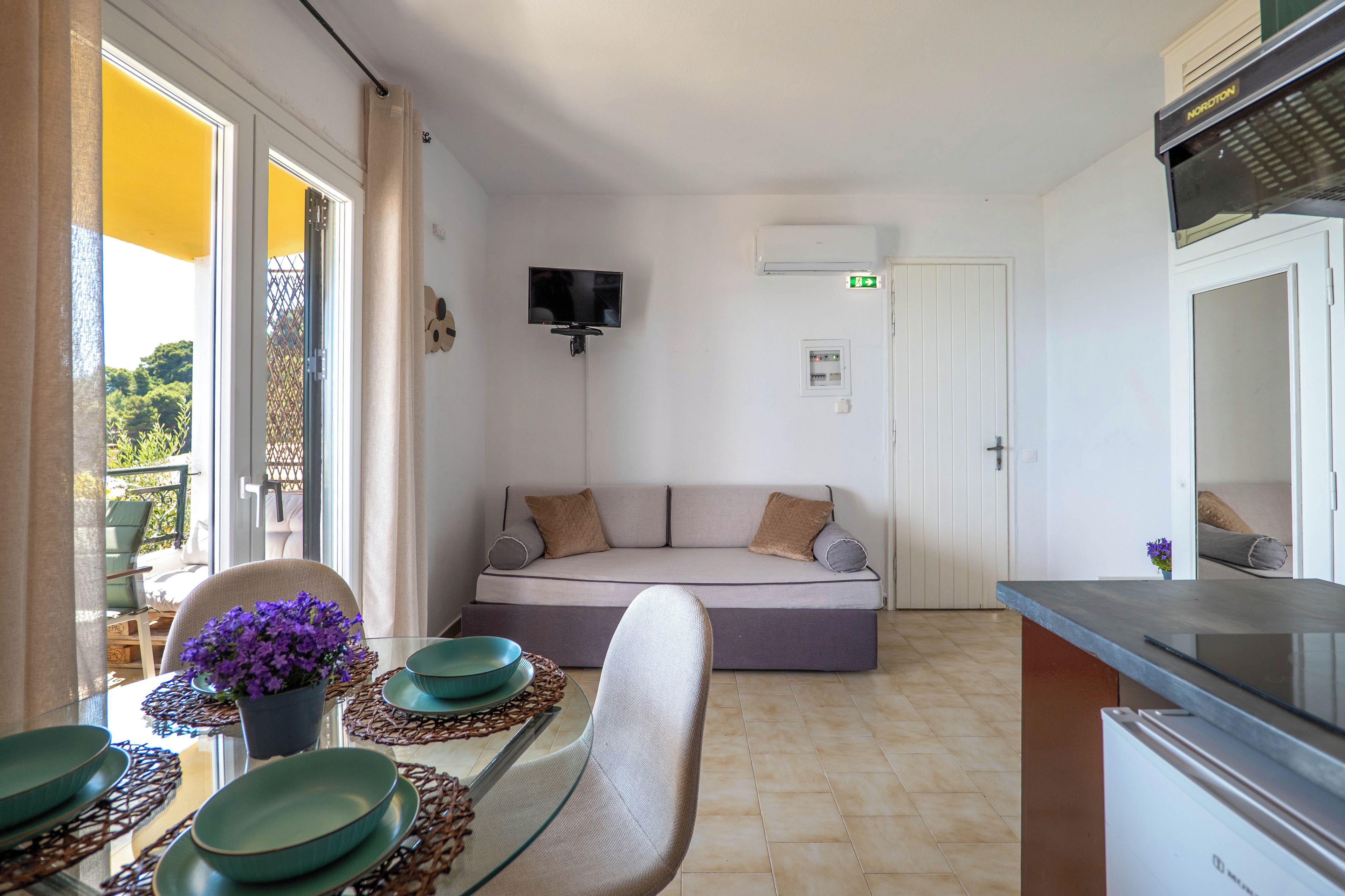 A modern living room with a sofa, a dining table with chairs, a flat-screen TV mounted on the wall, and a kitchen area with a countertop.