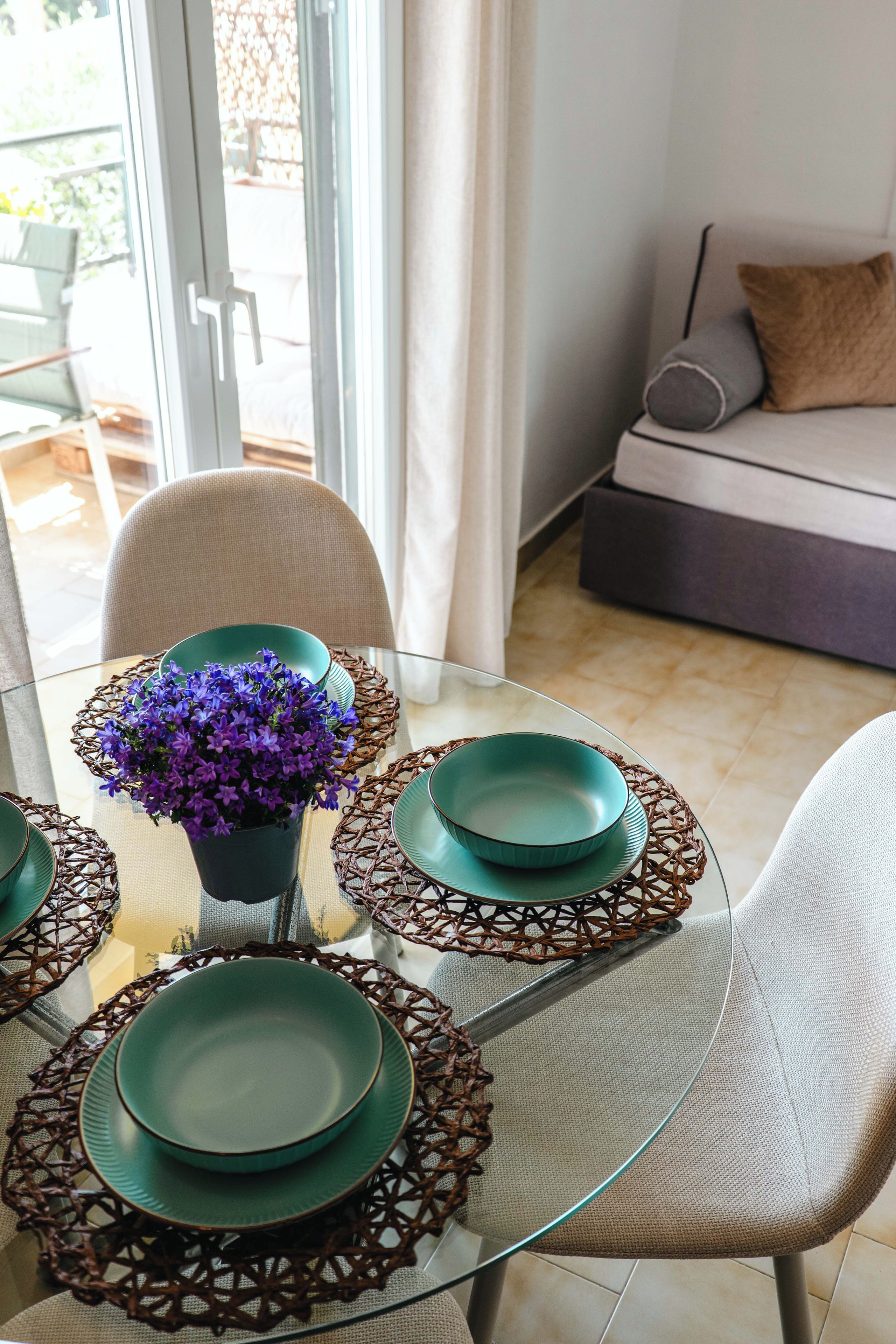 A dining area with a glass table, green plates, and a potted plant with purple flowers.